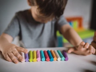 A little boy of three years sits at a table with colored crayons and plasticine