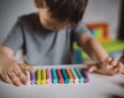 A little boy of three years sits at a table with colored crayons and plasticine
